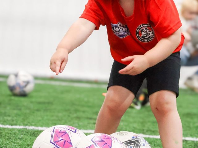 A young girl in a red First Kicks shirt stepping over a row of pink footballs