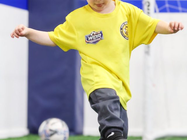 A toddler in a yellow HTFC Bumblebees shirt swinging a foot through a pink ball