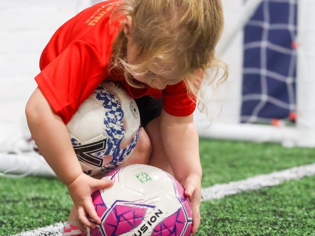 A young girl in a red First Kicks shirt crouching to gather up two footballs