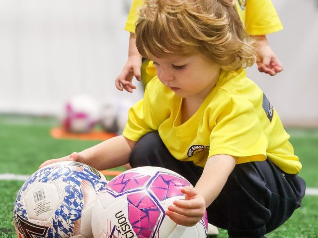 A toddler in a yellow First Kicks shirt crouching between two footballs in a marker ring