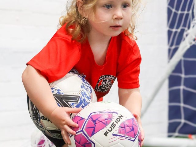 A young girl in a red BAFC Badgers shirt picking up two footballs at once