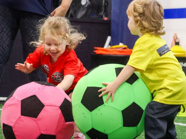 Two children laughing as they dive onto giant pink and green footballs