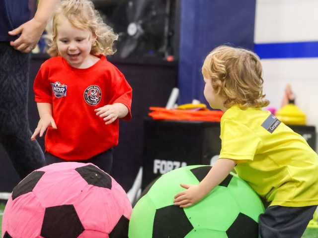 Two children pushing giant pink and green footballs across the indoor pitch