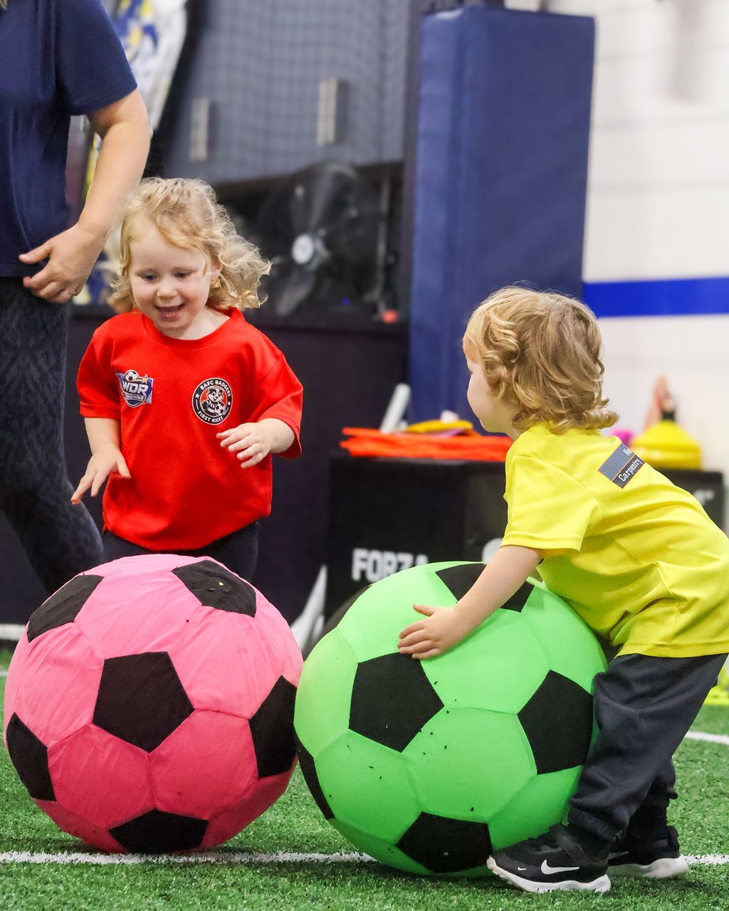 Two children pushing giant pink and green footballs across the indoor pitch