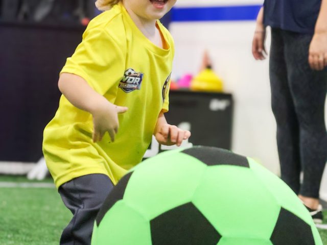 A boy in a yellow First Kicks shirt laughing as he chases a giant green football