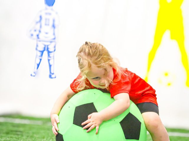A child in red hugging a giant green football, the painted player figures on the arena wall behind