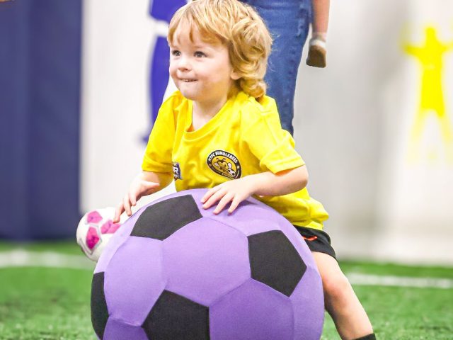 A boy in a yellow HTFC Bumblebees shirt leaning over a giant purple football, grinning