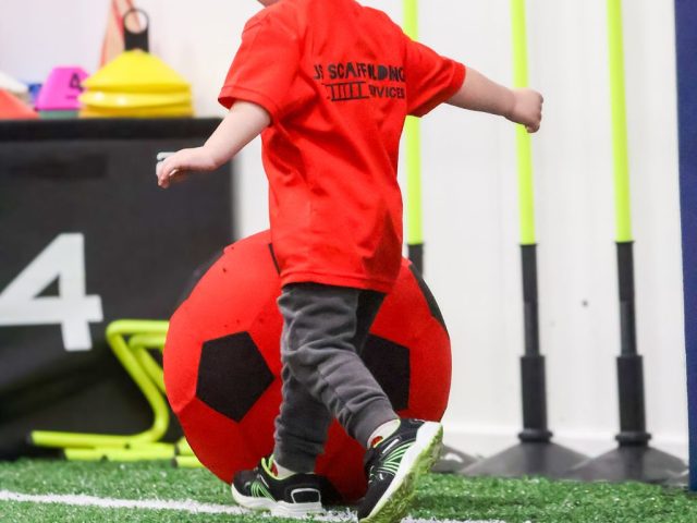 A child in a red JS Scaffolding Services shirt walking a giant red football along the line