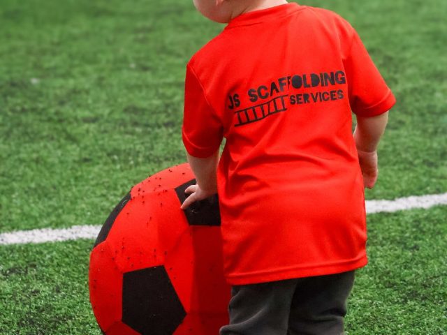 A child in a red JS Scaffolding Services First Kicks shirt steadying a giant red football
