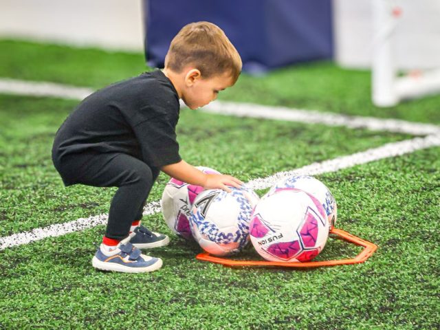 A young player in black crouching to gather footballs into a marker ring