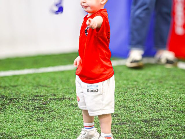 A young player in a red top standing with a foot on the ball, pointing across the pitch