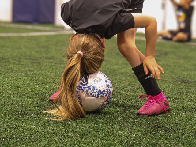 A young player bent right over a ball, ponytail on the astro and pink boots planted
