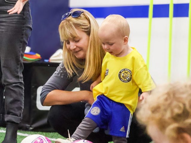 A parent crouching beside her toddler in a yellow First Kicks shirt, pink footballs set out in front of them