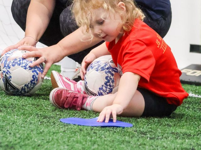 A parent and a toddler in a red First Kicks shirt pressing a blue marker onto the astro together