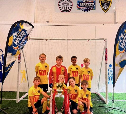 A Harborough Town junior team posed with a trophy in front of the goal, between the WDR feather flags