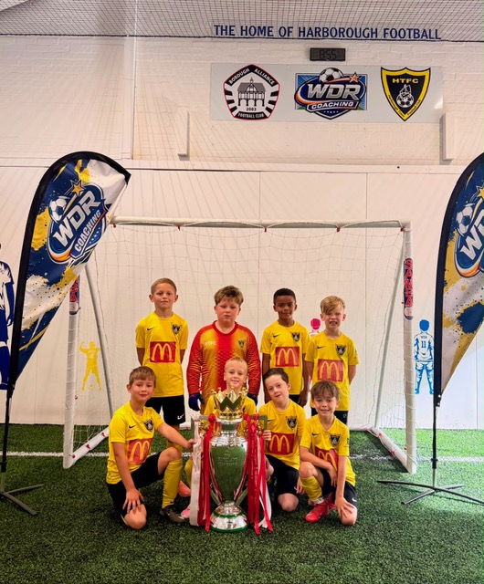 Eight children in yellow and red kit posing with the Premier League trophy in front of a goal at the WDR Football Centre, under the gable lettered “The home of Harborough football”
