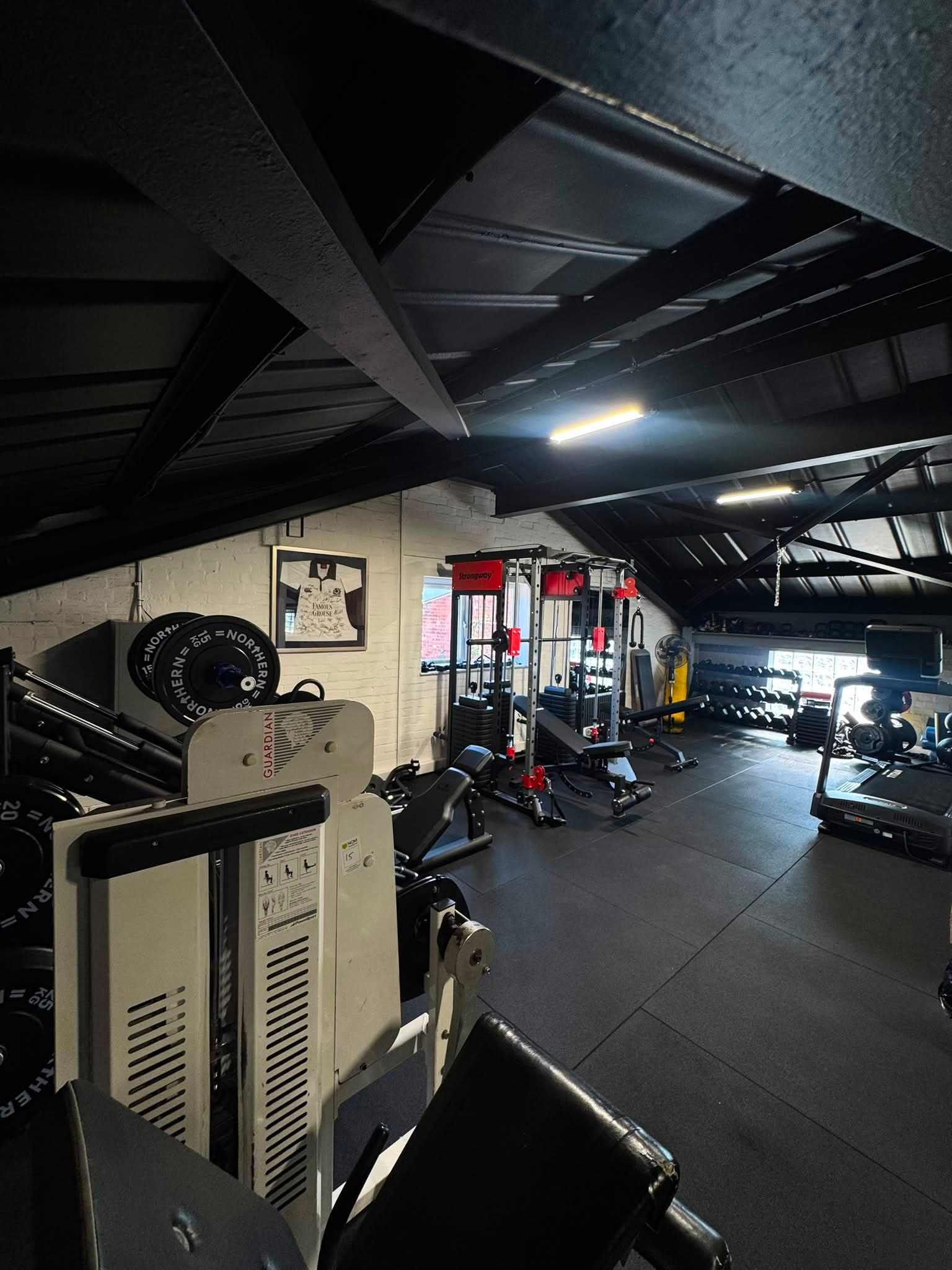 The studio gym looking down the room — leg press and weight plates in the foreground, squat rack, benches and dumbbells beyond, under a black pitched roof