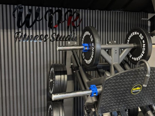 The WDR Fitness Studio signage on the slatted wall of the gym, with a barbell rack and weight plates in front of it