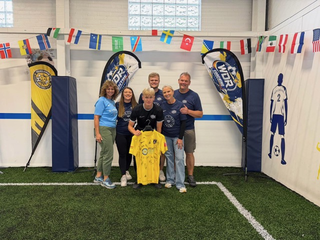 The WDR Football Centre team together on the astro pitch, holding a signed Harborough Town shirt, with the WDR feather flags behind them