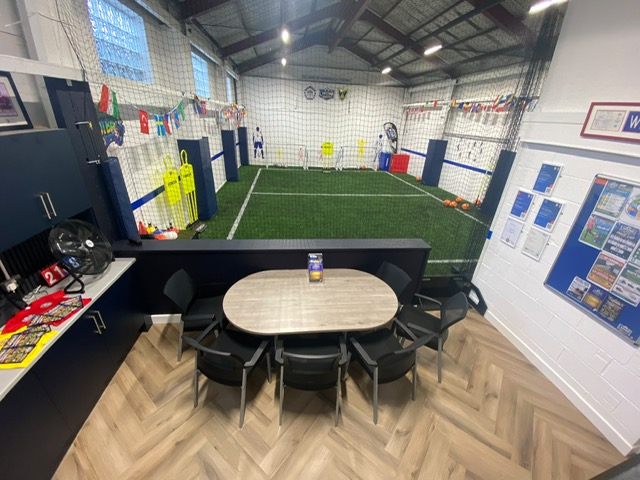 The seating area at the WDR Football Centre — a table and chairs on a herringbone floor, looking out over the indoor astro pitch through the netting